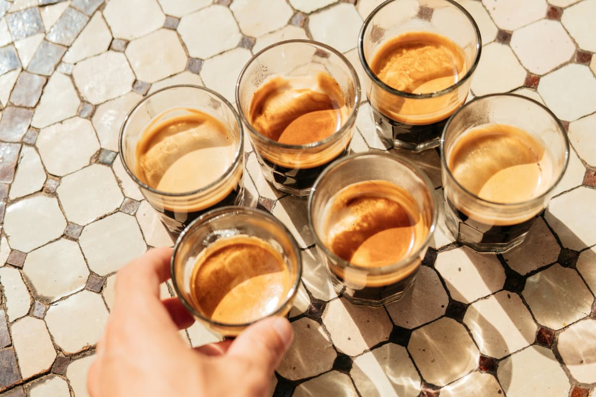 Overhead view of six glasses of espresso arranged on a decorative octagonal tiled surface in warm sunlight, with a hand reaching in from the lower left to pick up the nearest glass.